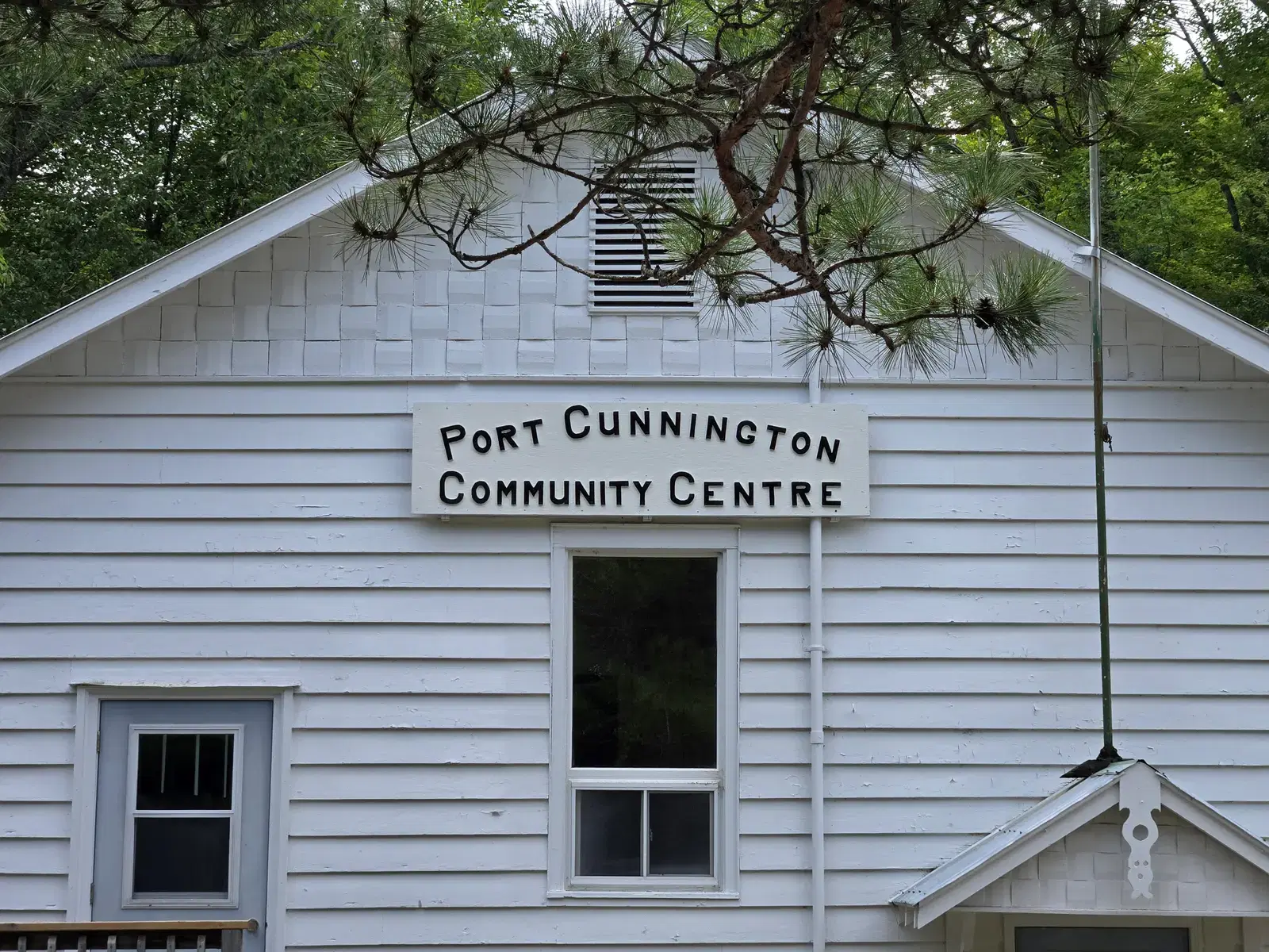 The shingled gable end of the hall with its hand-lettered sign, framed by the branch of a white pine.
