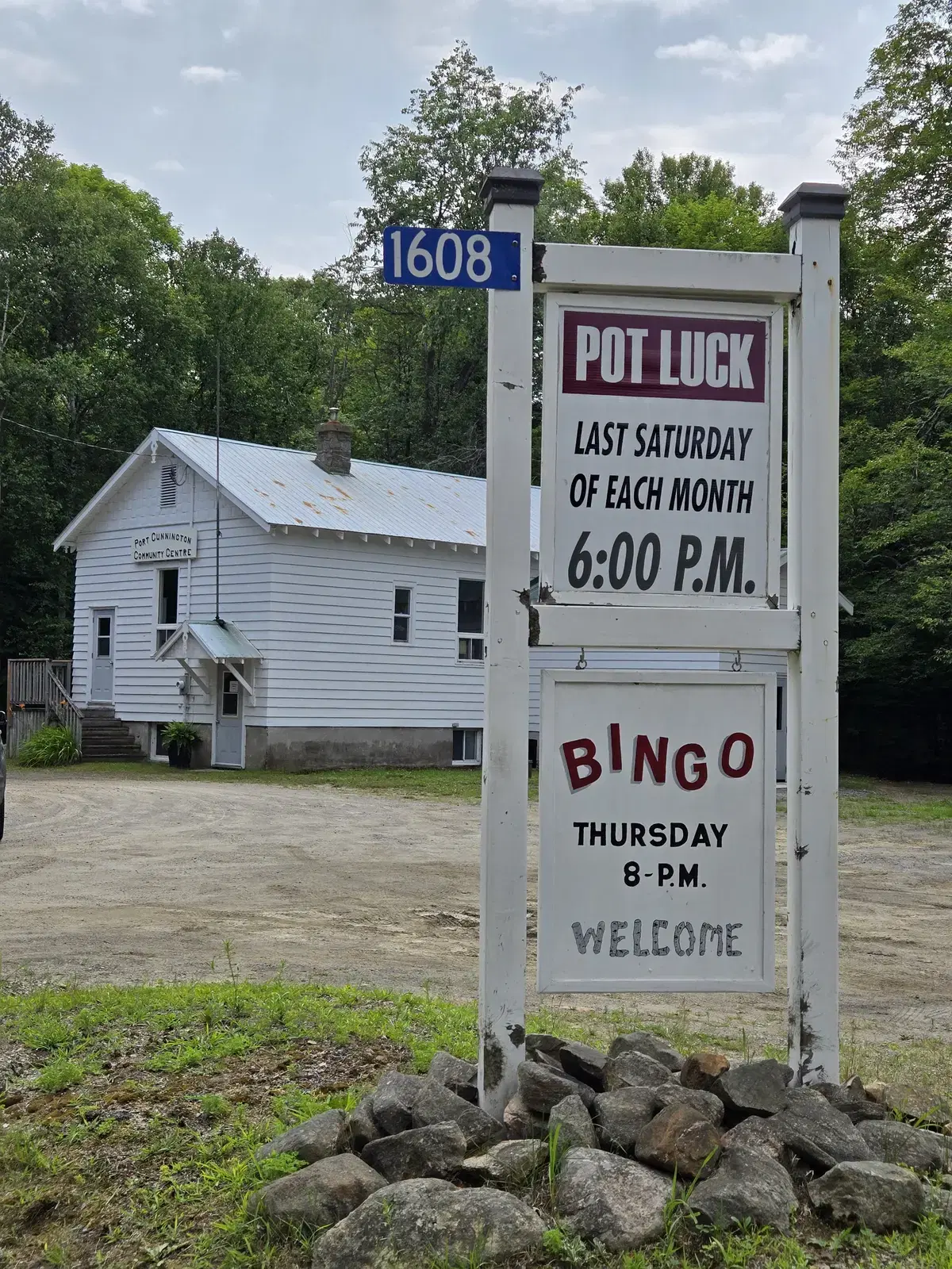 The white roadside noticeboard on its posts, lettered 'Pot Luck, last Saturday of each month, 6:00 P.M.' above 'Bingo, Thursday 8 P.M. Welcome', with the civic number 1608 on a blue plate and the hall behind.